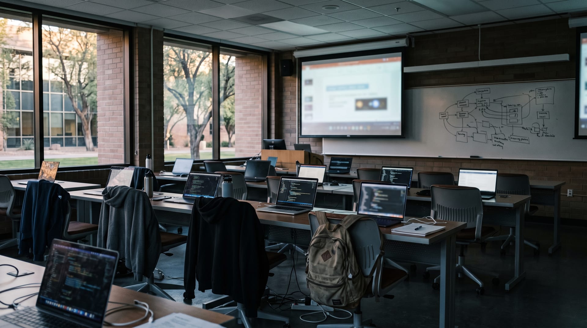 A university computer lab between workshop sessions: rows of desks with open laptops, hoodies and backpacks left on chairs, a projector screen with a blurred slide, and a whiteboard with a hand-drawn learning-path diagram.