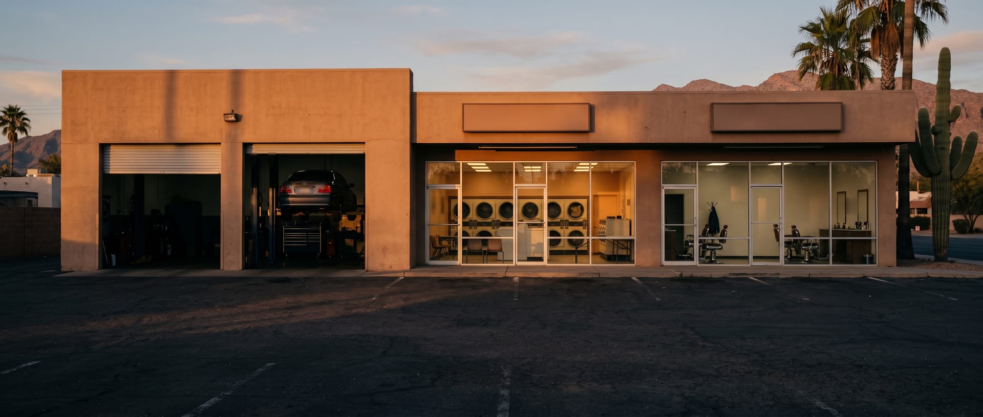A row of small service businesses in Phoenix at golden hour: an auto repair shop with open bay doors, a laundromat and a barbershop, under palm trees.