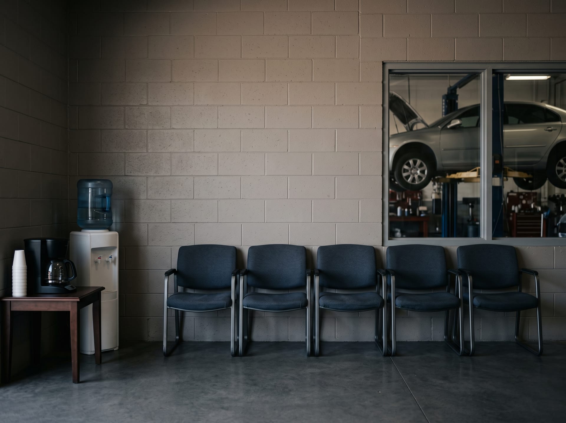 The waiting area of an auto repair shop seen straight on: a row of chairs under a bare stretch of painted wall, a coffee station, and a window into the service bay.