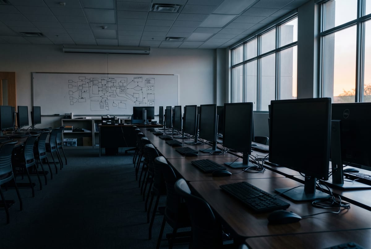 An empty university computer lab at dawn, rows of monitors dark, a whiteboard with a system diagram at the front.